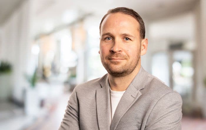 Ein junger, freundlich schauender Mann, in einem hellgrauen Sakko und einem weissen Shirt.