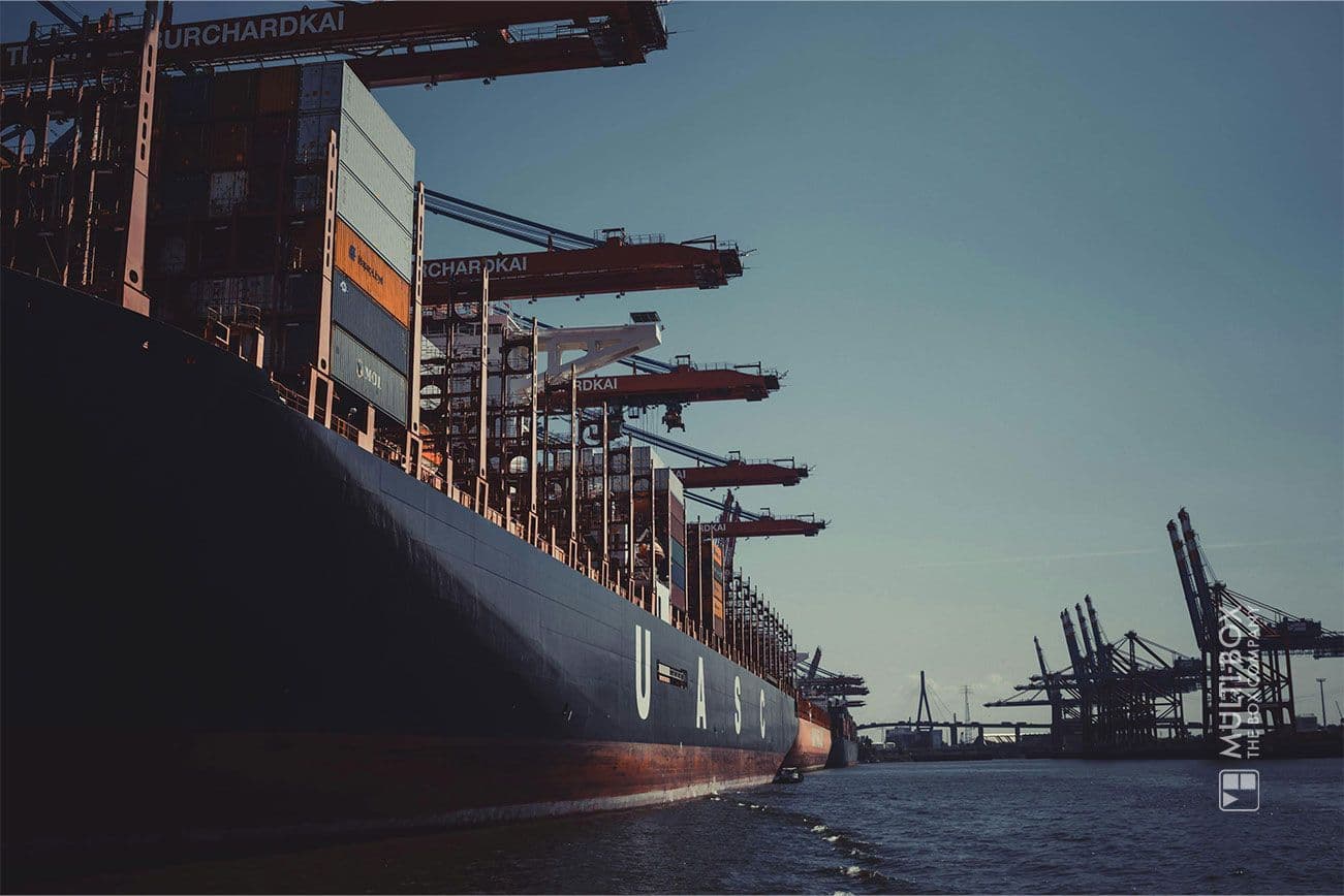 A container ship at the port, photographed at sunset. The focus is on the cranes and containers.