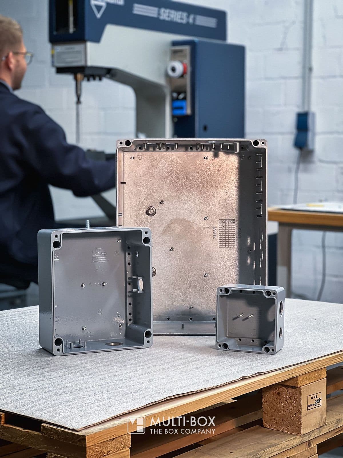 An employee works on machines for processing enclosures in the background, while various enclosure parts are laid out on a table in the foreground.